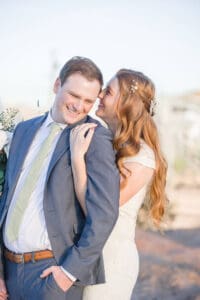 A bride whispers in her groom's ear while standing and laughing in a garden at their cactus joe's wedding