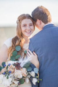 A bride hugs onto her groom's arm while standing in a desert at sunset