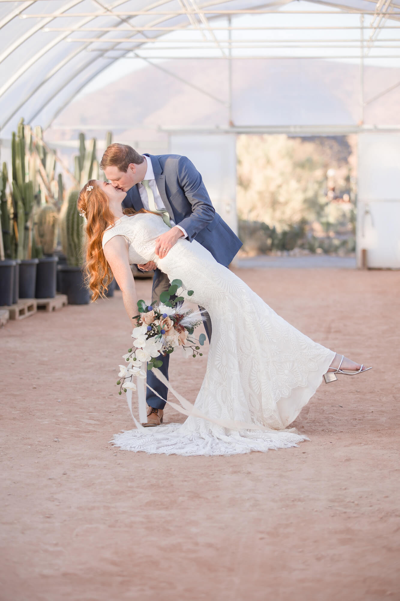 A groom in a blue suit dips his bride for a kiss while standing in a greenhouse