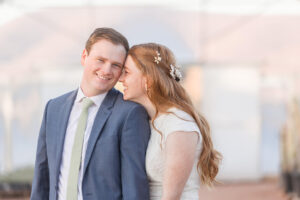 A bride snuggles onto her groom's shoulder while they stand in a greenhouse