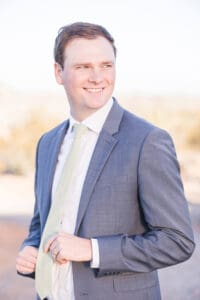 A groom adjusts his green tie while standing in a desert garden in a blue suit