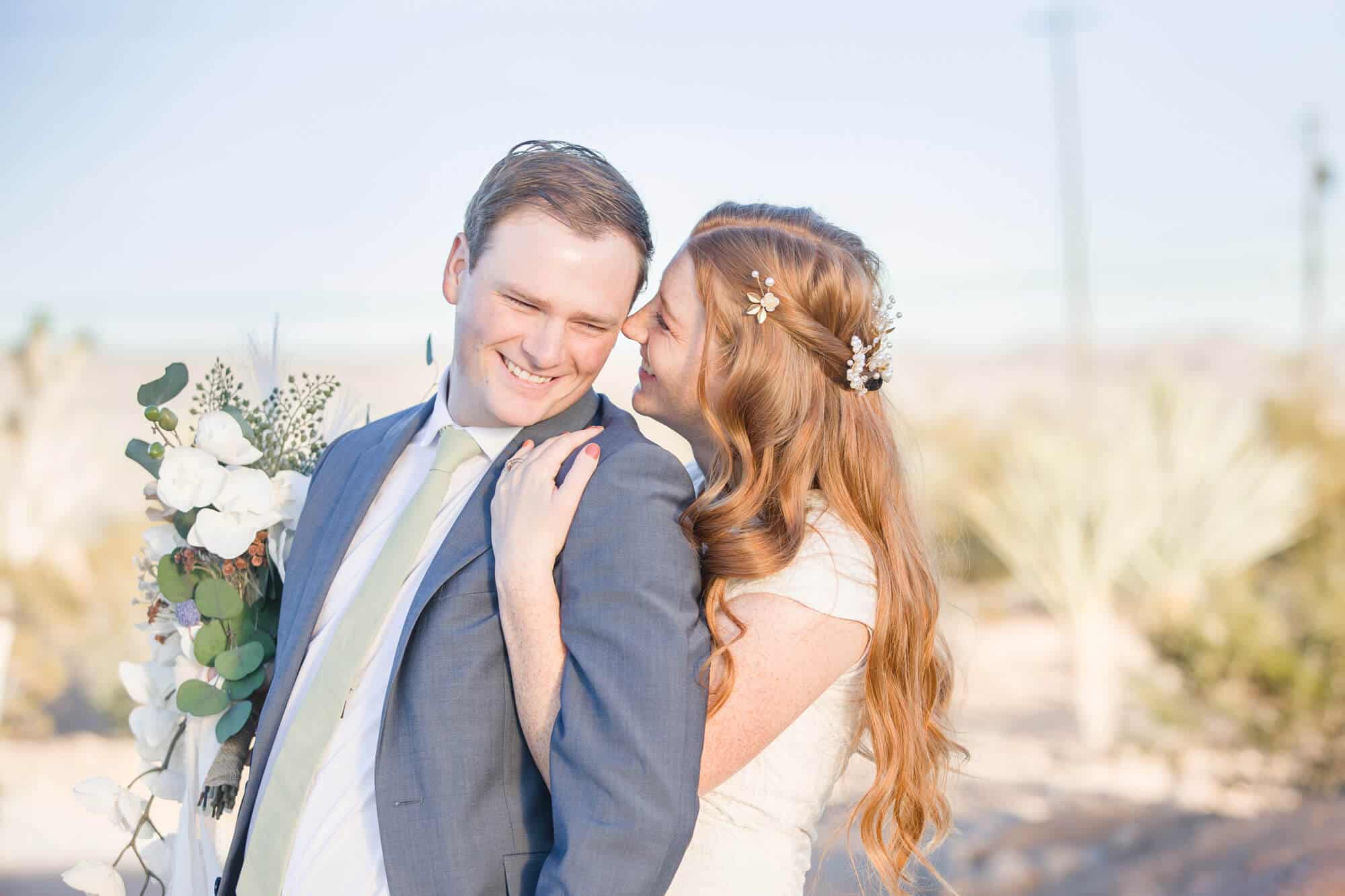 A bride hugs her groom in a blue suit from behind and whispers into his ear