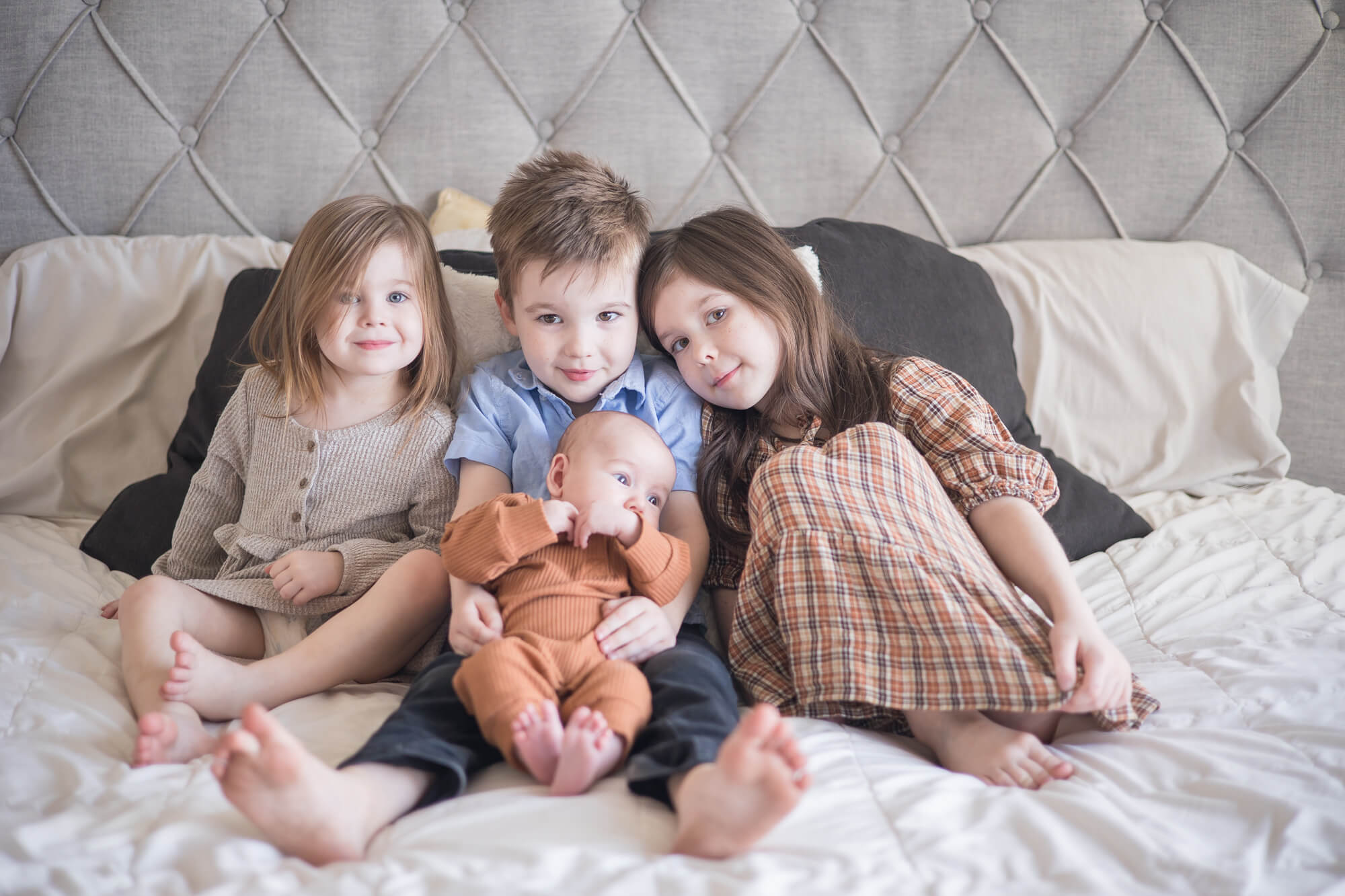 Three toddler siblings sit on a bed with their newborn baby brother in the boy's lap