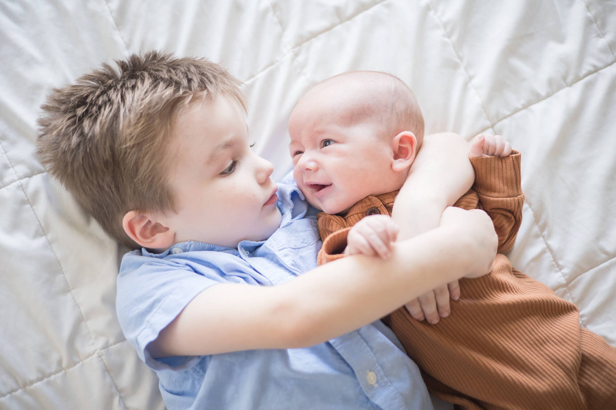 A toddler boy in a blue shirt hugs his newborn baby brother while laying on a white bed