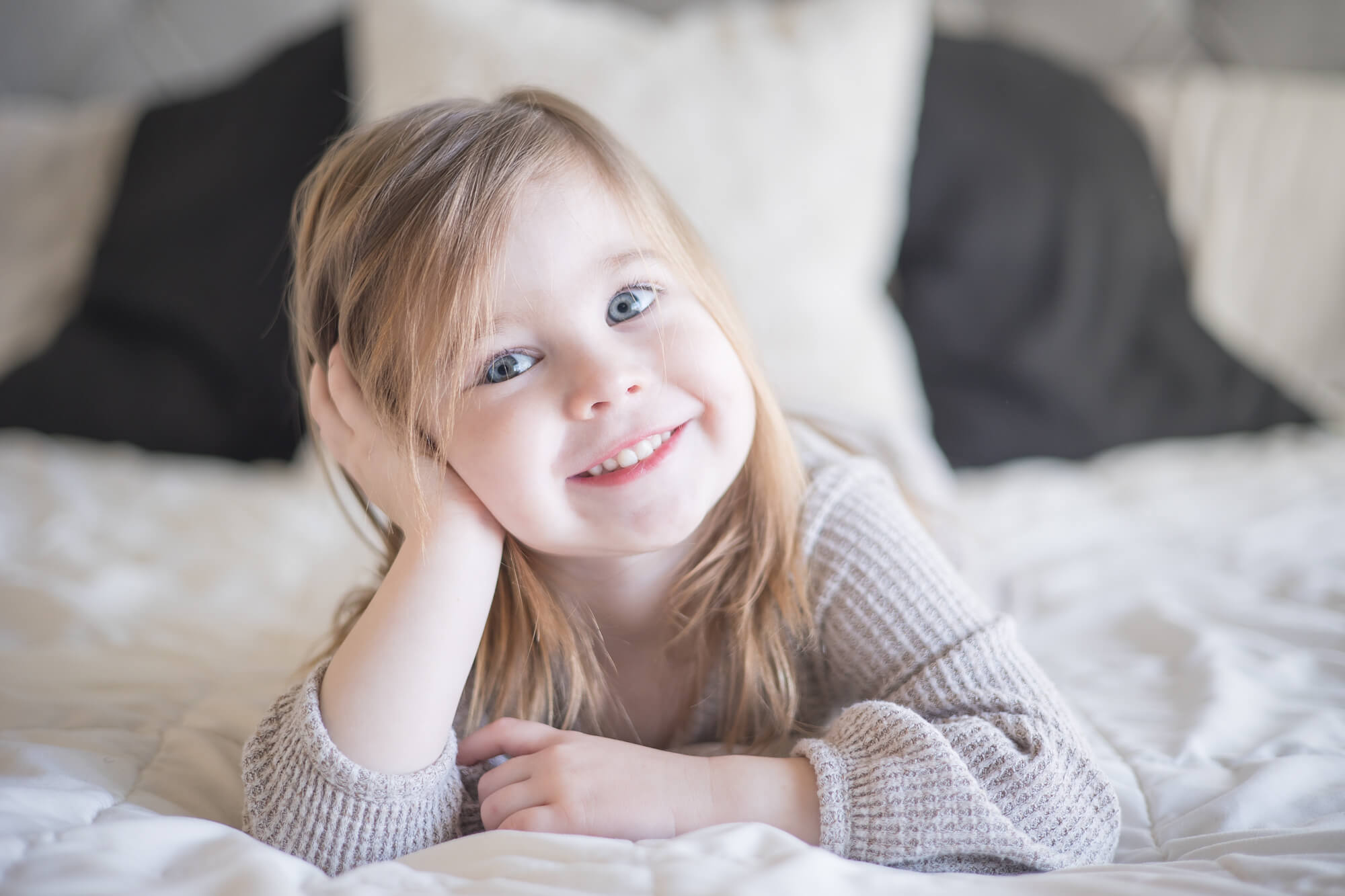 A toddler girl lays on a bed under a window resting her hand on her head