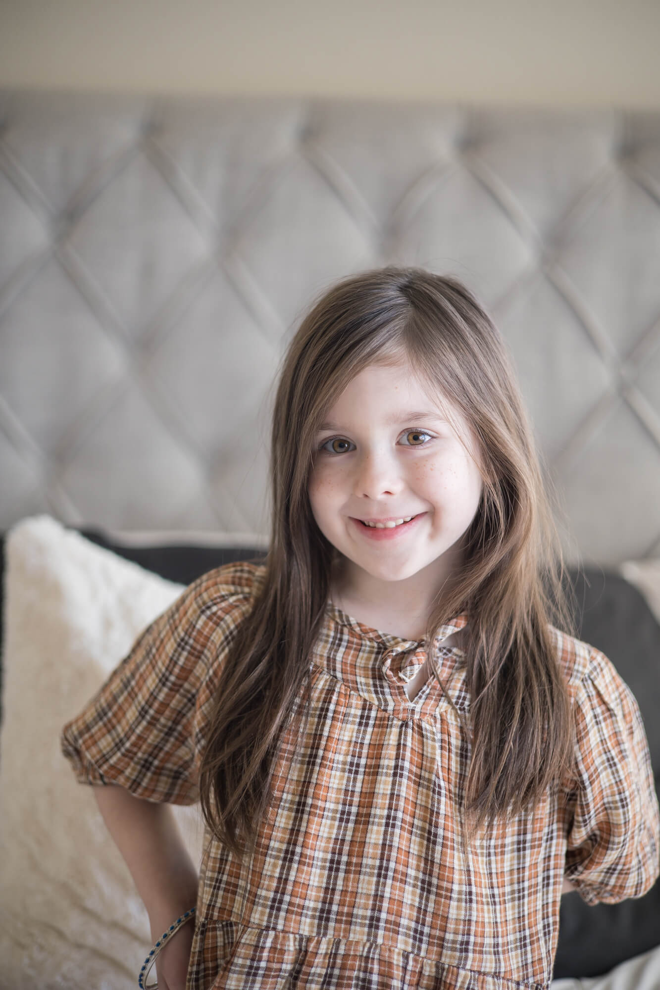 A toddler girl in a plaid dress sits on a bed smiling before meeting las vegas nannies