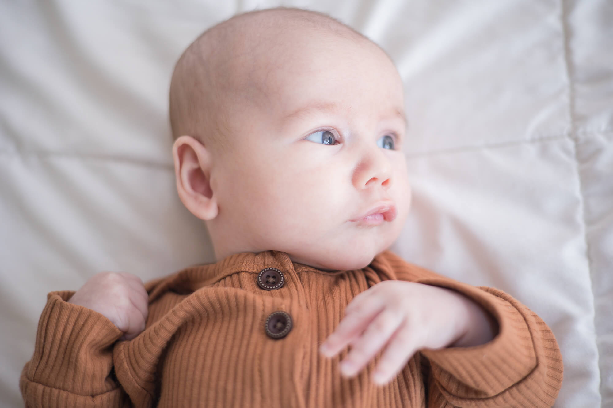 A newborn baby lays on a white bed looking out a window in a brown onesie before meeting las vegas nannies