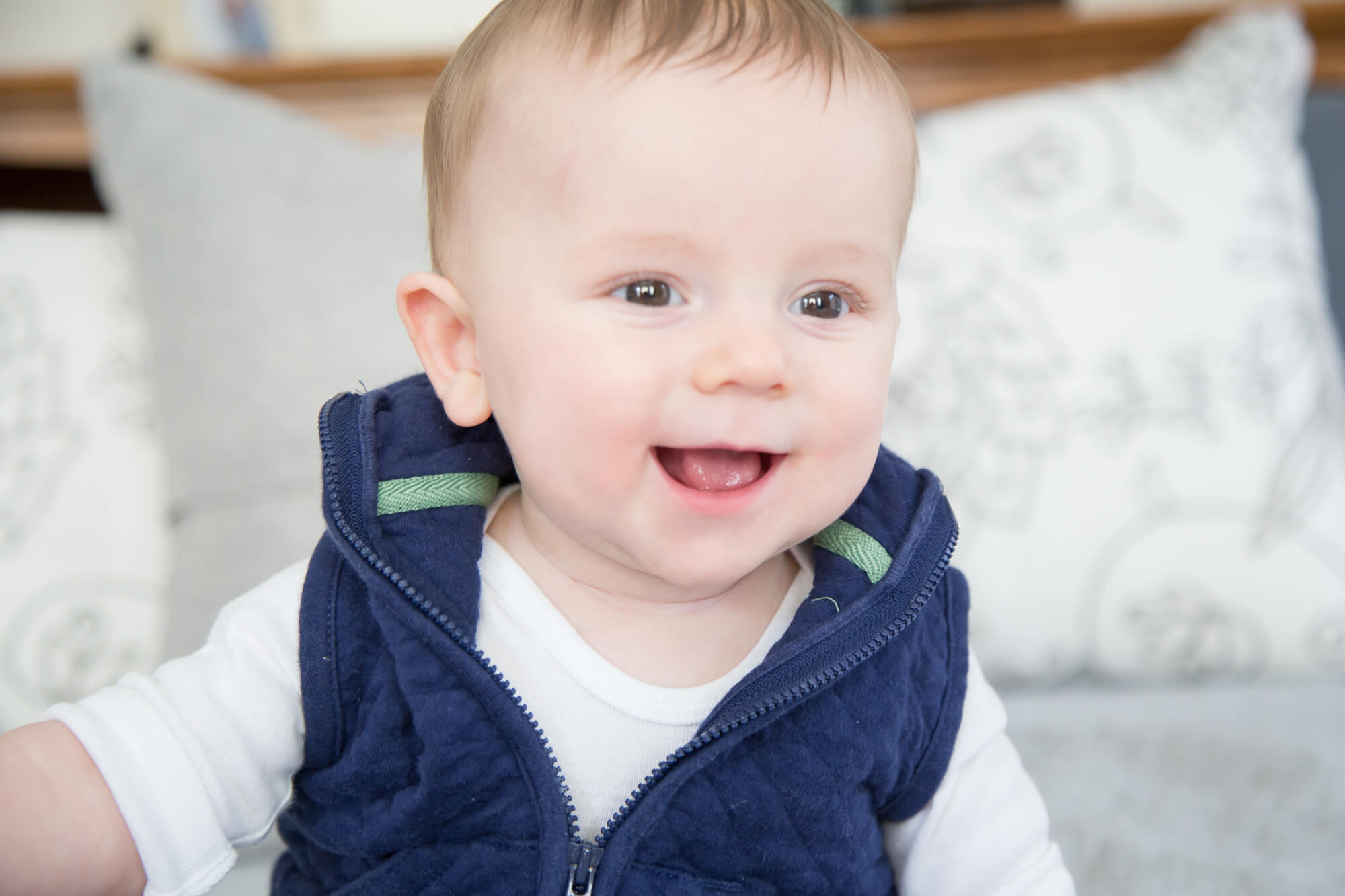 A toddler boy smiles big while playing on a bed in a blue vest