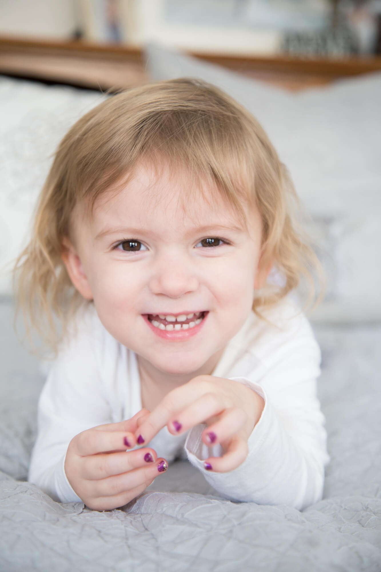 A toddler girl with purple nails giggles while laying on a bed