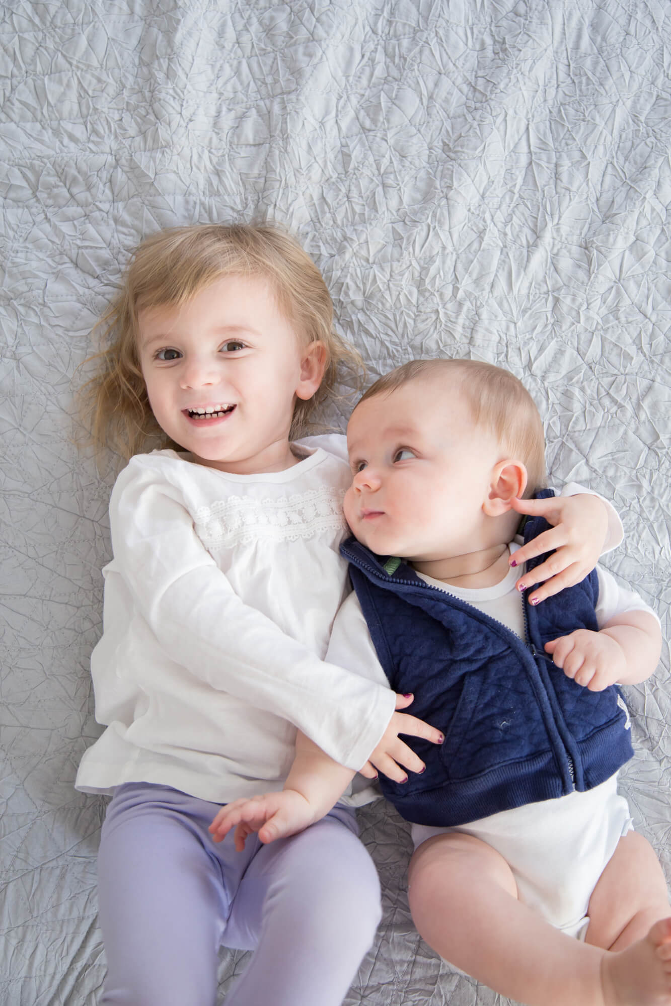 A toddler girl lays on a bed hugging her infant baby brother in a blue vest before visiting las vegas pediatrician