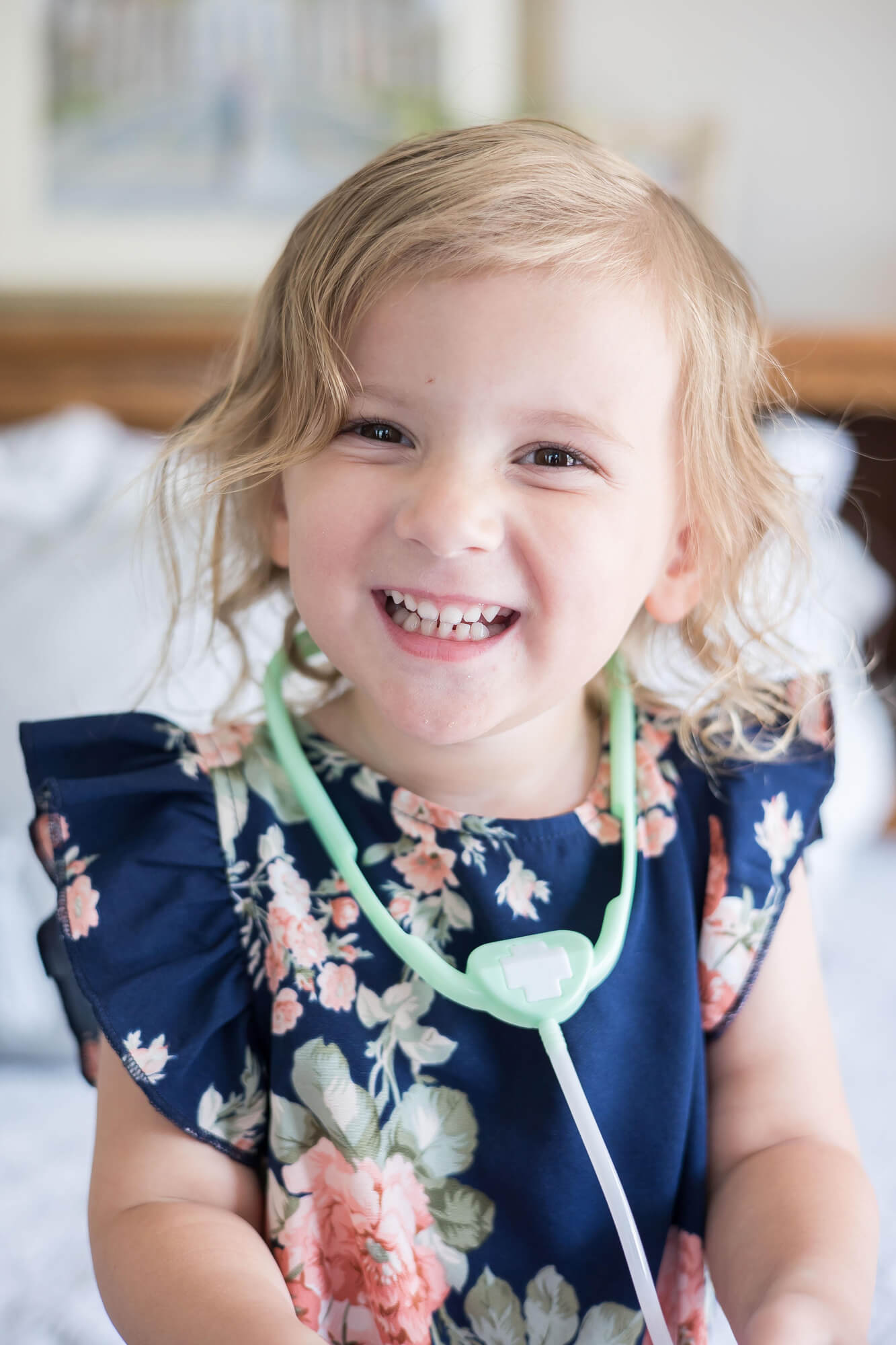 A toddler blonde girl smiles and laughs while wearing a toy doctor's tool on a bed