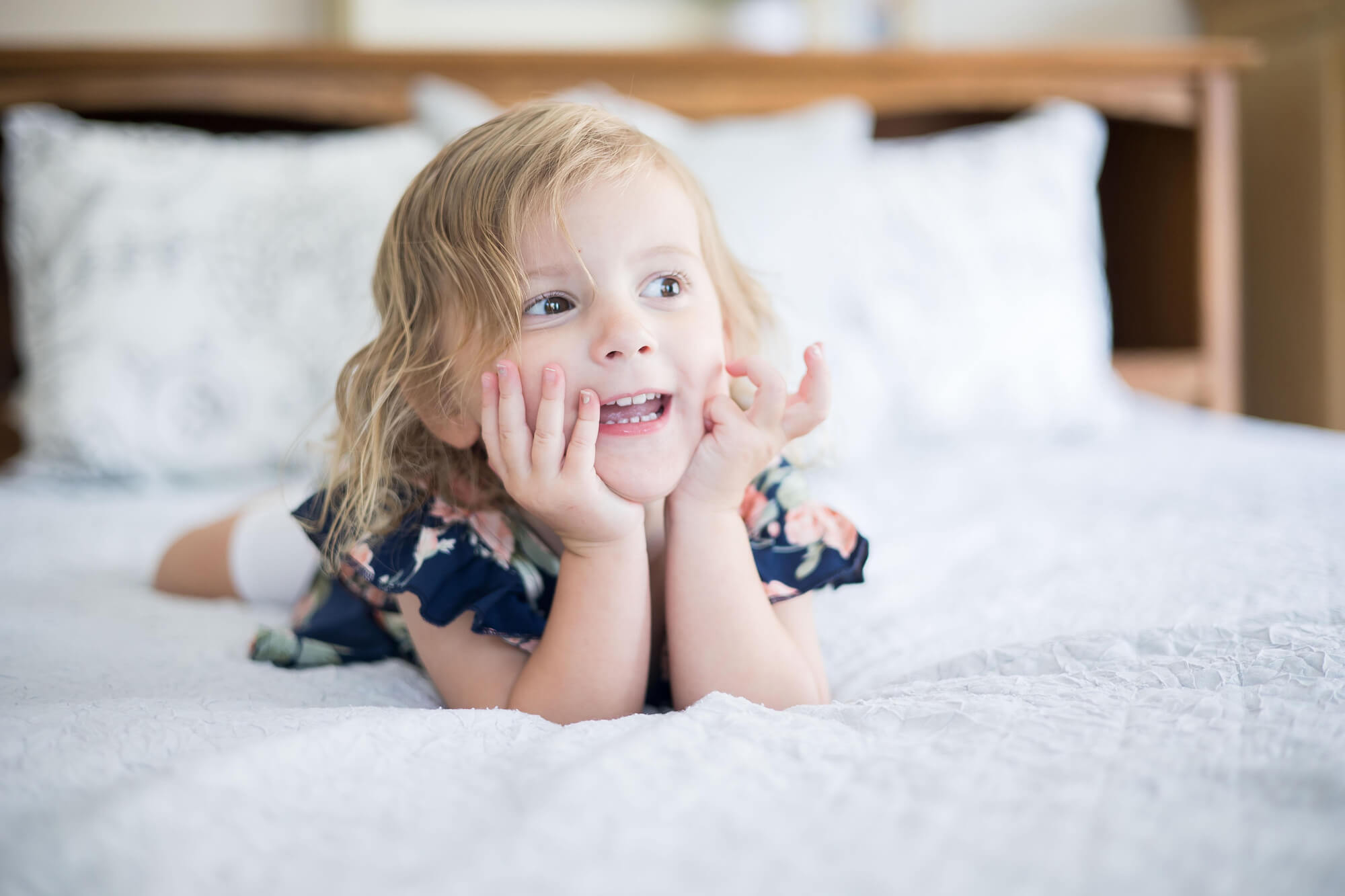 A toddler girl in a blue floral print dress rests her chin in her hands while talking on a bed before visiting las vegas preschools