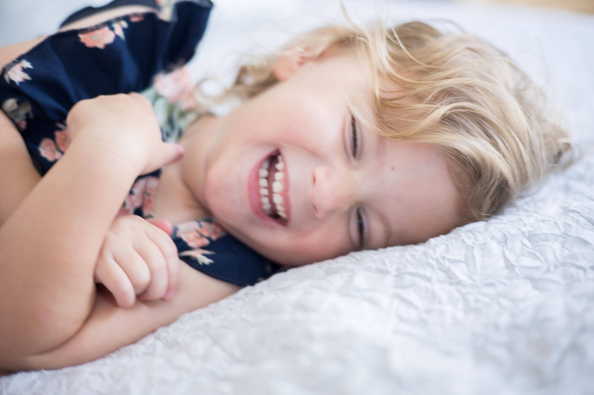 A toddler girl laughs and giggles while laying on a white bed in a blue dress