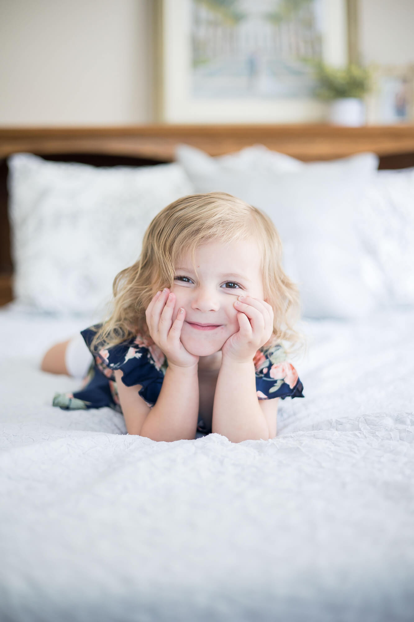 A toddler girl lays across a white bed in a blue floral print dress resting her head in her hands before visiting las vegas preschools