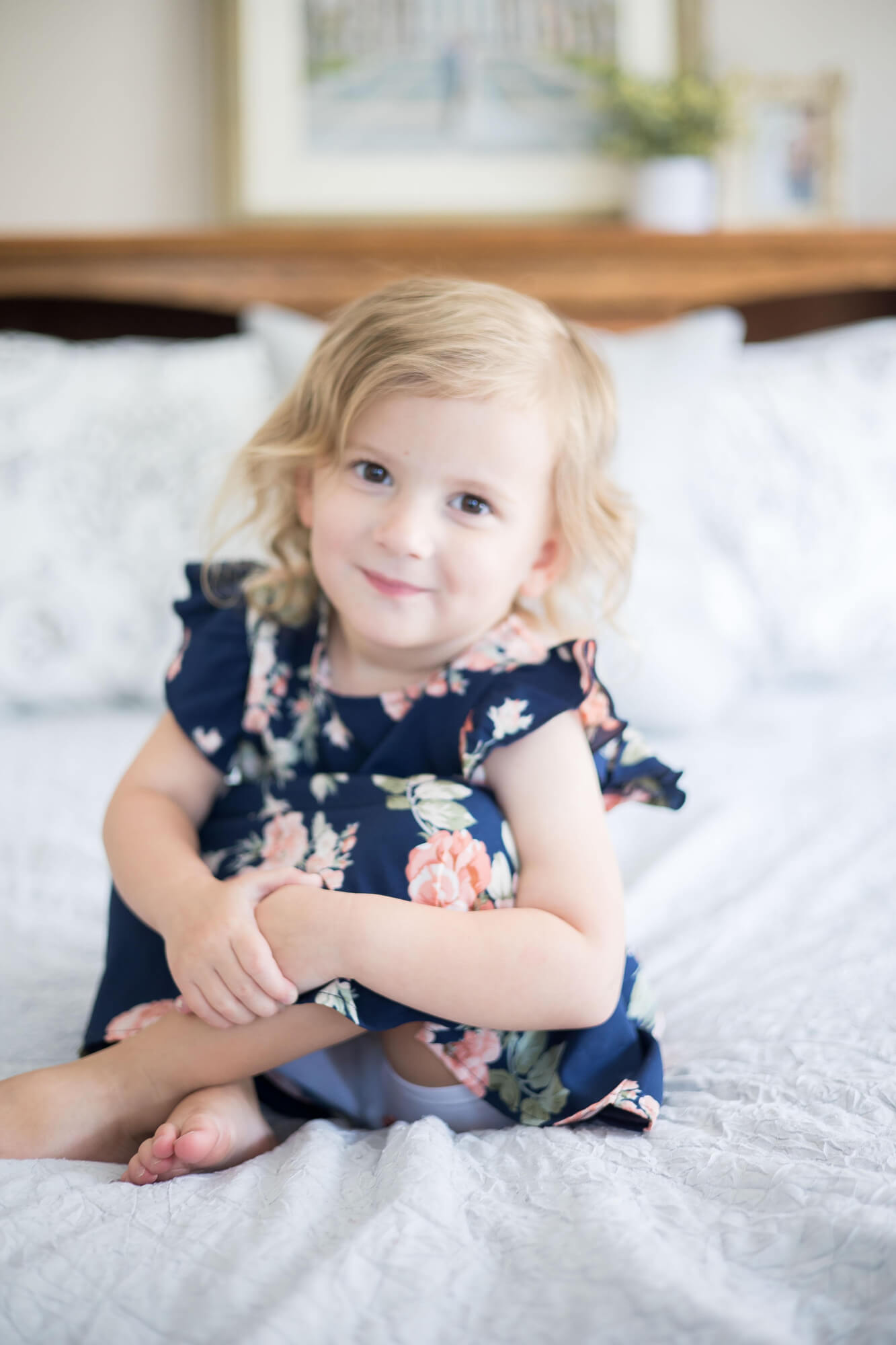 A toddler girl in a blue floral print dress sits on a white bed smiling before visiting las vegas preschools