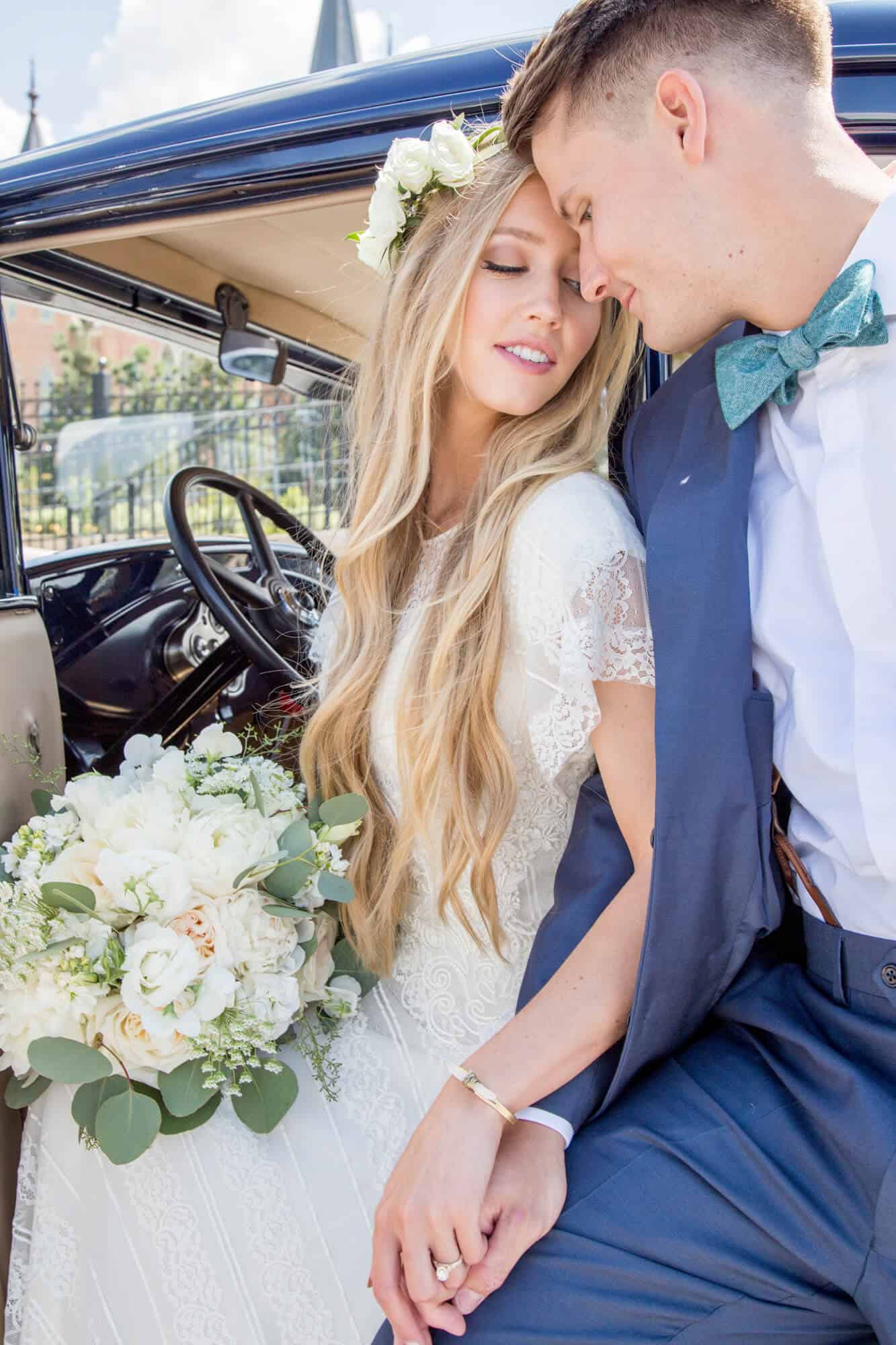 A bride and groom sit on the edge of a vintage car holding hands and leaning in for a kiss