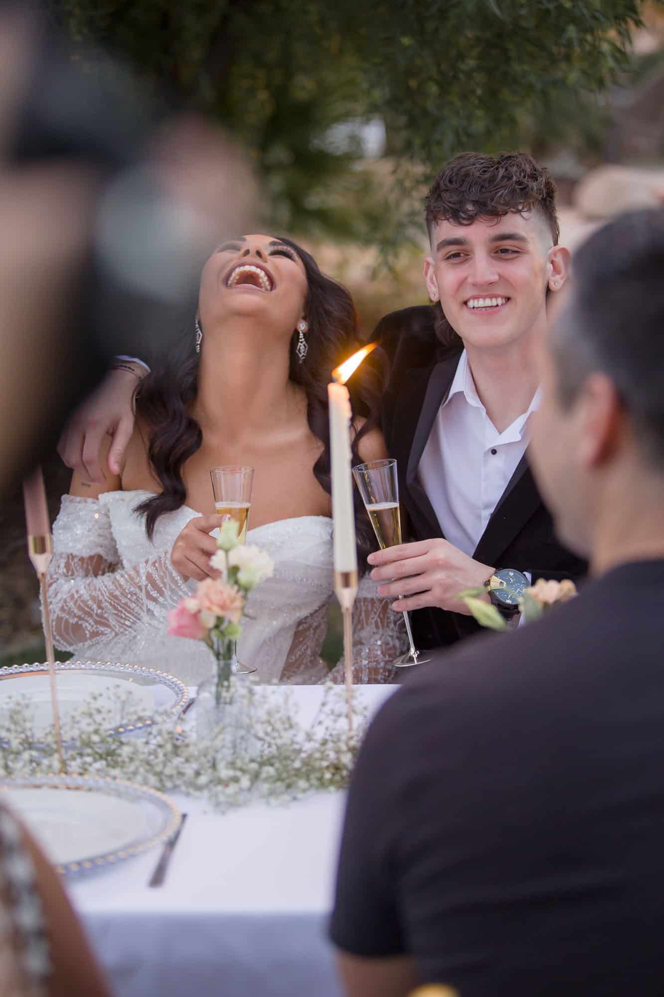 Newlyweds sit at a reception table laughing and holding champagne at one of the outdoor wedding venues las vegas