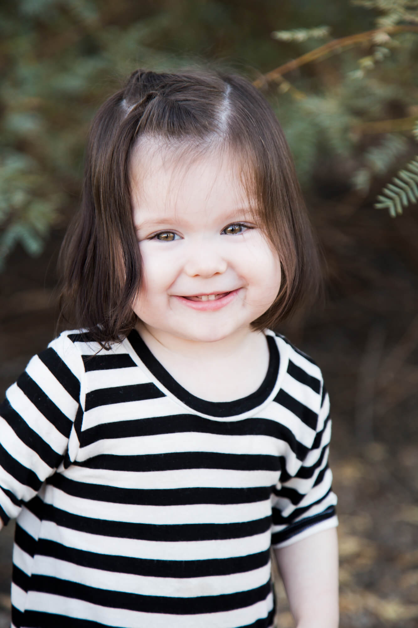 A toddler girl smiles big while exploring some woods in a striped black and white dress